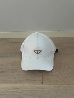 Vit keps från PR i syntet - Snygg vit keps från PR med klassisk böjd skärm och justerbar rem bak. Kepsen har Pradas ikoniska metallogo framtill och är tillverkad i ett glansigt syntetmaterial som ger en clean och modern look.