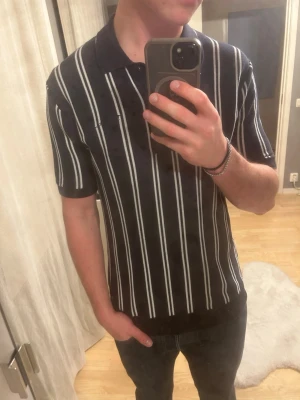 Svart kortärmad skjorta med ränder - Snygg svart skjorta med vita vertikala ränder och korta ärmar. Skjortan har klassisk krage och ribbade muddar vid ärmslut och nederkant. Perfekt för en stilren och modern look. Materialet är bomull och är väldigt bekvämt. 