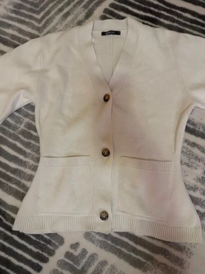 Vit kofta med knappar och fickor - Bara använd 1 gång. Tyvärr är tröjan för stor.             Från Gina tricot. Stilren vit kofta med v-ringning och tre stora knappar framtill. Koftan har två praktiska fickor och ribbade muddar vid ärmslut och nederkant. Perfekt att slänga över en t-shirt för en enkel och clean look.