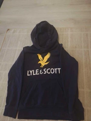 Mörkblå hoodie från Lyle & Scott - Snygg mörkblå hoodie från Lyle & Scott med stor gul logga och text på bröstet. Klassisk huva och magficka framtill. Perfekt för en avslappnad och trendig stil. Tillverkad i mjukt material som känns skönt mot huden. Storleken är 8 till 9 år gammal!!