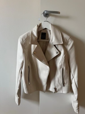 Ljusbeige skinnjacka med dragkedjor - Snygg ljusbeige skinnjacka med klassisk bikerkrage och silvriga dragkedjor framtill. Jackan har två fickor med dragkedja och markerade sömmar för en cool look. Perfekt för dig som vill ha en stilren och trendig jacka till din outfit.