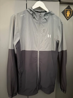 Grå vindjacka från Under Armour - Snygg tvåfärgad vindjacka från Under Armour i ljus- och mörkgrått. Jackan har huva, hel dragkedja, bröstficka med dragkedja och två sidofickor. Tillverkad i lätt och vindtätt material, perfekt för blåsiga dagar.