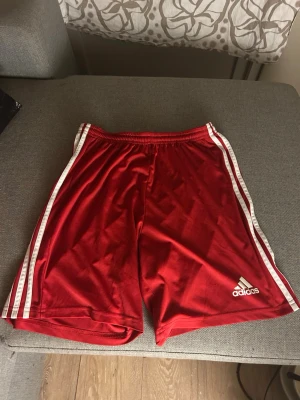 Röda Adidas shorts med vita ränder - Snygga röda shorts från Adidas med klassiska vita ränder längs sidorna och Adidas-logga på ena benet. Tillverkade i ett lätt och sportigt material med resår i midjan för skön passform. Perfekta för träning eller chill.