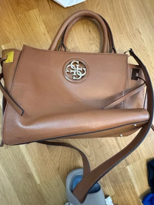 Brun GUESS väska  - Snygg brun handväska i konstläder med metallogo framtill. Väskan har både handtag och axelrem, samt flera fack inuti för smart förvaring. Perfekt för dig som vill ha en stilren och praktisk accessoar till vardagslooken.