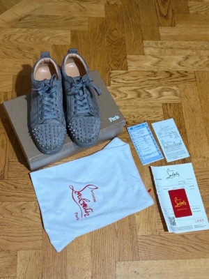 Gråa Louboutin sneakers med nitar - Snygga gråa sneakers från Christian Louboutin i mocka med coola silverfärgade nitar på tån och klassisk röd sula. Skorna har snörning och en låg profil som ger en clean look. Perfekta för dig som vill sticka ut med exklusiva detaljer.
