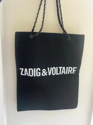 Svart shoppingväska Zadig & Voltaire - Snygg svart shoppingväska från Zadig & Voltaire med vit logga framtill. Väskan har kedjehandtag i metall som ger en cool touch. Perfekt för dig som vill ha en stilren och rymlig tote till vardagen. Materialet ser ut att vara tyg med en mjuk känsla. Väskans del där kedjan sitter är lite trasig, vilket jag gjorde en lätt lösning till som jag tycker fungerar. Men det är även väldigt lätt att laga 