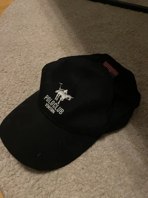 Svart keps Polo Club Vinson - Snygg svart keps från Polo Club Vinson med broderad logga framtill i vitt och justerbart spänne bak. Klassisk böjd skärm och mjukt material som sitter skönt på huvudet. Passar perfekt till en avslappnad streetwear-look.