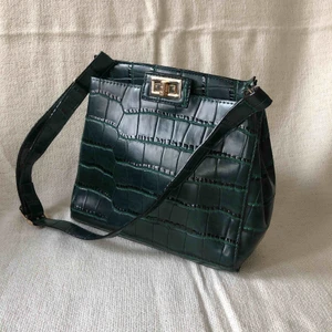  - Mörkgrön krokodilmönstrad väska med gulddetaljer. Fack på vardera sida av väskan. Slitage vid spännena av bandet därav priset.   Hämtas upp i Stockholm eller köpare står för frakt.