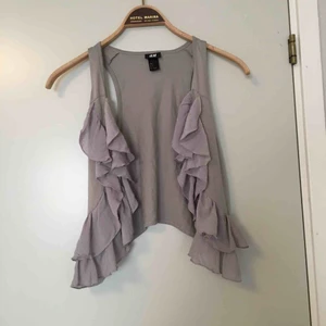  - Sommrig väst med tunna volanger, i gråa toner. Passar perfekt om man vill göra en tshirt eller klänning mer festlig eller över en skjorta! 