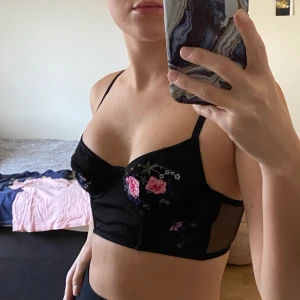 Svart BH/top med broderade detaljer! - Superfin svart BH/top med rosa/blå broderade blomsterdetaljer. Meshliknande material. Snygg att ha under en djupt ringad kavaj! Från topshop i storlek 70b/32b. Har reglerbara band & 3 olika lägen på knäppet i ryggen. Frakt ingår!