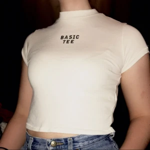  - Polo T-shirt med trycket "basic tee". Tröja är benvit och slutar runt naveln beroende på kroppsform