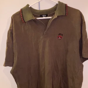 Vintage Stüssy T-shirt - Säljer denna coola vintage Stüssy pikéen från 90-talet!! Den är i en brun/grön mörk färg i storlek L men det är möjligt att den är större än så. Eftersom att den är gammal är den i ett litet slitet skick då kragen har blivit noppig och det saknas en knapp, annars är skicket bra!! Köparen står för frakt 🤎💚