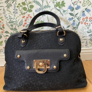 DKNY väska - Snygg väska från DKNY. Bra med plats utan att kännas för stor. Guldiga detaljer, det klassiska DKNY tyget. Ett stort fack samt två mindre med dragkedja. Även ett kortfack samt det lilla facket på utsidan. Fler bilder kan skickas vid önskemål. 