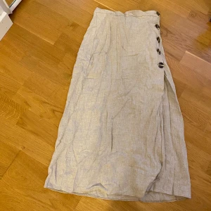 Kjol med knappdetalj - Säljer en lång beige kjol med knappdetaljer samt en snygg slits. Rätt mycket trådar vid slitsen men det är inget man ser när man har på den. Är osäker men tror materialet är i linne ev en linne mix. Är 167 och den är bra i längden. Köpt på asos
