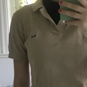 Beige Piké från Lacoste  - Beige poke från lacoste. Supersnygg men kommer tyvärr inte längre till användning. Tröjan har inga hål men en fläck som går att se på bild två. Fläcken var dock inget jag själv sätt innan. Köpt second hand och sedan dess endast använd 1-2 gånger. Det står att storleken är 3 men jag skulle uppskatta att storleken är en s som även passar xs. Den första kan man se hur tröjan ser ut på mig som är en storlek s.