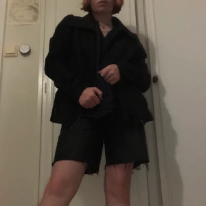 Svart jacka med dragkedja - En svart jacka med dragkedja och stor krage. Dragkedjor längs handlederna och fickor på sidorna. Resår på ryggen för en närsittande plagg. L är den storleken den är menad att ha men den passar mer som en M.