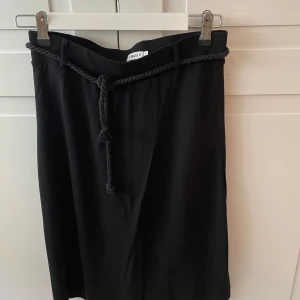 Filippa K svart kjol - Svart kjol från Filippa K. Kjolen har en omlott slitts framtill. Separat bälte i tyg ingår. Varan är i fint skick. 