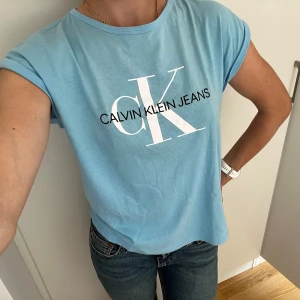 Ljusblå T-shirt från Calvin Klein - Ljusblå lös T-shirt i storlek 16 år/ S.  T-shirten har vikta ärmar och Calvin Klein Jeans tryck på bröstet