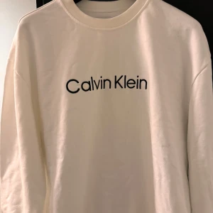 Vit tröja från Calvin Klein - En stilren vit tröja från Calvin Klein med svart text på framsidan. Tröjan har långa ärmar och en rund halsringning.
