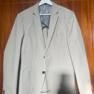 Beige kostym från Lindbergh White - En elegant beige kostym från Lindbergh White i storlek herr 48. Kostymen är slim fit och består av en kavaj med två knappar och matchande byxor. Kavajen har ett blommigt innerfoder och byxorna har en klassisk design med hällor för bälte. Kan skickas eller mötas upp i Umeåområdet. 