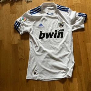 Vit fotbollströja från Adidas - En vit fotbollströja från Adidas med blå detaljer. Tröjan har korta ärmar och en krage. På framsidan finns Real Madrids klubbmärke och sponsorlogga 'bwin.com'. På baksidan står det 'REAL MADRID' under kragen.