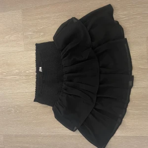 Svart volangkjol - Säljer en superfin svart volangkjol. Den är kort och har en elastisk midja som gör den bekväm att bära! Kjolen är gjord av ett lätt och luftigt material. Helt oanvänd!