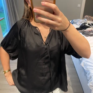 Svart blus från H&M - Säljer en elegant svart blus från H&M. Blusen har en lös passform och korta ärmar. Den är tillverkad i ett glansigt material som ger en lyxig känsla. Perfekt för både vardag och fest!