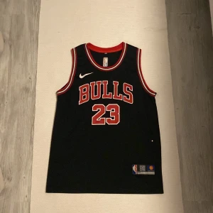 Svart Chicago Bulls basketlinne - Säljer ett svart Chicago Bulls basketlinne från Nike. Linnet har nummer 23 och namnet Jordan på ryggen. Det är i nyskick och har röda och vita detaljer runt halsen och ärmhålen. Perfekt för basketfans eller som en cool streetwear-stil. Köp direkt eller ge prisförslag 🏀