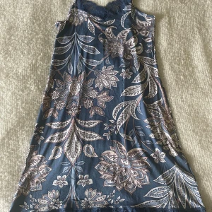 Blommig klänning med spetsdetaljer - Ett vacker nattlinne med blommigt mönster i blå och vita nyanser. Nattlinnet har spetsdetaljer vid halsen och är ärmlös.