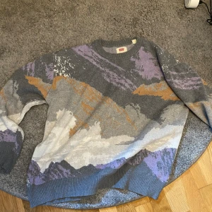 Mönstrad stickad tröja från Levi's - Säljer en supercool stickad tröja från Levi's i storlek M. Tröjan har ett unikt mönster i grått, lila, vitt och orange som ger den en riktigt snygg look. Den är perfekt för höst och vinter och är riktigt mysig att ha på sig. Tröjan har långa ärmar och en normal passform. Perfekt för att hålla sig varm och stilren under de kallare månaderna!