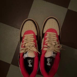 BAPE STA sneakers i beige, brun och röd - Säljer ett par college dropout bapesta sneakers i beige, brun och röd färg som är designade av Kanye west. Skorna är använda en gång och har inga defekter. Skorna har en stjärndetalj på sidan och är tillverkade i skinn. De har snörning och en röd yttersula som ger en cool kontrast. Perfekta för både vardag och fest!