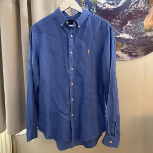 Säljer en snygg blå linneskjorta från Polo Ralph Lauren i storlek L. Skjortan är slim fit och har långa ärmar. Den är tillverkad i ett bekvämt material och har knappar framtill. Perfekt för både vardag och fest!