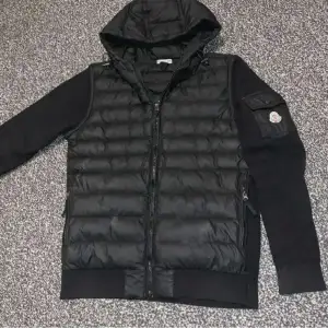 Säljer en snygg svart jacka från Moncler. Jackan har en quiltad design på framsidan och ärmarna är av ett annat material. Den har en huva och dragkedja framtill. På vänster ärm finns en ficka med Moncler-loggan. Perfekt för höst och vår!