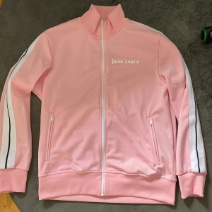 Rosa träningsjacka från Palm Angels - Säljer en snygg rosa träningsjacka från Palm Angels i skick 9/10 som är använd fåtal gånger. Jackan har en dragkedja framtill och två fickor med dragkedjor. Den har vita och svarta ränder längs ärmarna och en hög krage. Perfekt stilren vardagsjacka! 