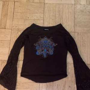 Tröja med mesh-ärmar  - Otrolig brun tröja med vida mesh-ärmar. Coolt tryck på framsidan med blå-glittriga detaljer. Ärmarna har små blommiga mönster.   Storlek S-M   
