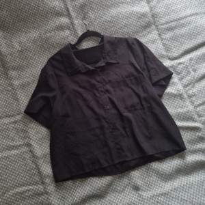 En svart kortärmad skjorta, använd någon enstaka gång. En bröstficka och detalj i ryggen. Avslappnad passform. Tunn polyester, ej stretchig.