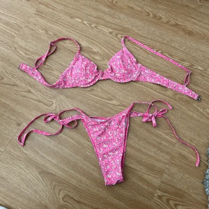 Rosa blommig bikini  - Rosa bikini med vita blommor. Bygel-bh med uttagbara vadderings ilägg och lågt skuren knytbar bikinitrosa. 