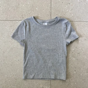 Grå ribbad kortare t-shirt/magtröja- GINA - Säljer denna söta ribbade tröjan/t-shirten från Gina i storlek S💘 Tröjan är i bra skick utan slitningar, hål eller fläckar och använd endast ett fåtal gånger!!