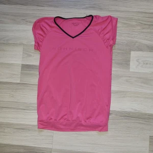 Rosa kortärmad topp från Röhnisch - En rosa kortärmad topp från Röhnisch med svart kant vid halsringningen. Toppen har en bekväm passform och är tillverkad i ett lätt och andningsbart material, perfekt för träning. storlek 150/160