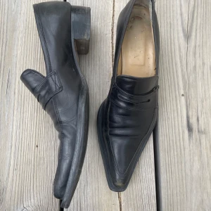 Spetsiga svarta loafers - Svarta skinnloafers med en spetsig tå och en 2 cm klack. Slitna i sulan och tån. Uppskattad storlek 37. Oklart märke men troligen Novita.