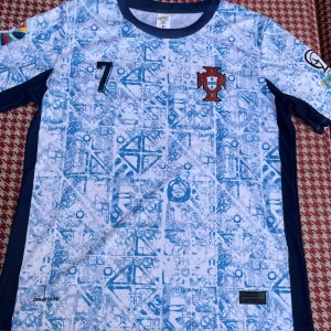 Vit och blå fotbollströja från Portugal - En vit och blå fotbollströja från Portugal med nummer 7 och namnet Ronaldo på ryggen. Tröjan har korta ärmar och ett geometriskt mönster i blått. Den har även Portugal-emblemet på bröstet och är tillverkad med Dri-FIT-teknologi.