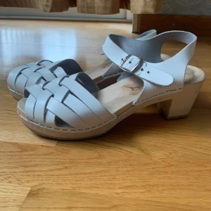 Ljusa sandaler med klack och spänne - Ett par ljusa sandaler med flätad design och justerbart spänne. Sandalerna har en klack och är i använt skick.