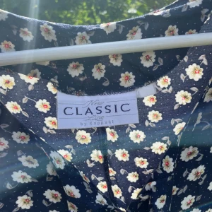 Figursydd mönstrad klänning  - Figursydd mörkblå blommig klänning med knappar fram och en rosett längst upp vid knapparna. Sval och fint fall. 100% viskos.