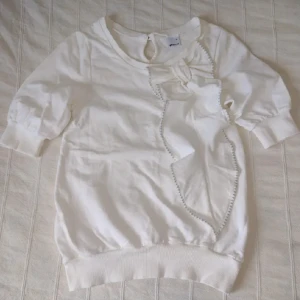 Vit tröja med volang, rosett och puffärm - storlek XS - Vit/benvit mjuk tröja med mudd nertill och i ärmsluten. Stor volang och rosett på framsidan. Puffärmar. Sweatshirtmaterial, 100% bomull. Använd en gång. Storlek XS.