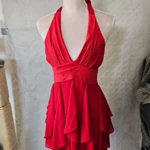 Röd klänning med volanger - En elegant röd klänning med volanger och halterneck-design. Klänningen har en omlott-detalj vid midjan och en v-ringad hals. Den är tillverkad av 100% polyester och har en dragkedja på baksidan.