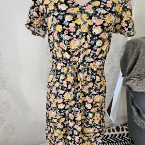 Blommig klänning från Vero Moda - En vacker blommig klänning från Vero Moda i storlek S. Klänningen har en svart bas med ett färgglatt blommönster i rosa, gult och blått. Den har korta ärmar och en v-ringad hals. Klänningen har en knytning i midjan för en smickrande passform.