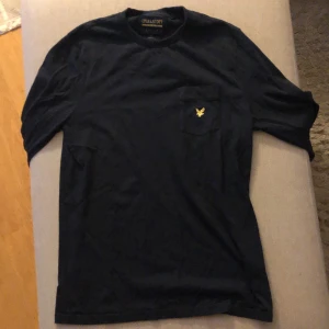 Mörkblå Tröja från Lyle & Scott - En Svart Tröja från Lyle & Scott med en broderad gul örn som är Lyle & Scotts logotyp. Tröjan har en relaxed fit och är kortärmad. Tröjan har Max använts 2 gånger och säljs därför lite dyrare. Tröjan är helt i nyskick!