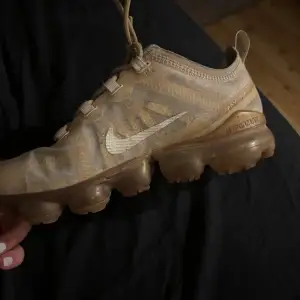 Ett par beige Nike Air Vapormax sneakers med snörning och genomskinlig sula. Skorna har en modern design med Nike-logotypen på sidan och är tillverkade i syntetmaterial.finbs ingwn sula i skon