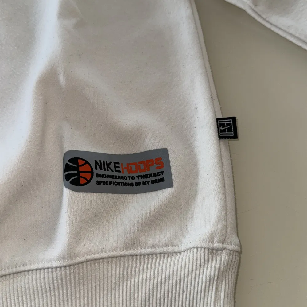 En vit sweatshirt från Nike med en röd basketboll och grå Nike-logotyp på bröstet. Tröjan har långa ärmar och ribbstickade muddar vid ärmslut och nederkant. Det finns även en etikett med texten 'Nike Hoops' på framsidan.. Tröjor & Koftor.