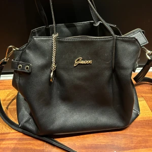 Guess Väska  - Den är en mellanstor väska som jag har älskat därav lite slitage. De slitage dom syns är på bandet, på kanten, ett litet brännmärke, lite läpppenna märjen i ett fack.  Mått: Bredd: 31cm Höjd: 28cm Djup: 14cm